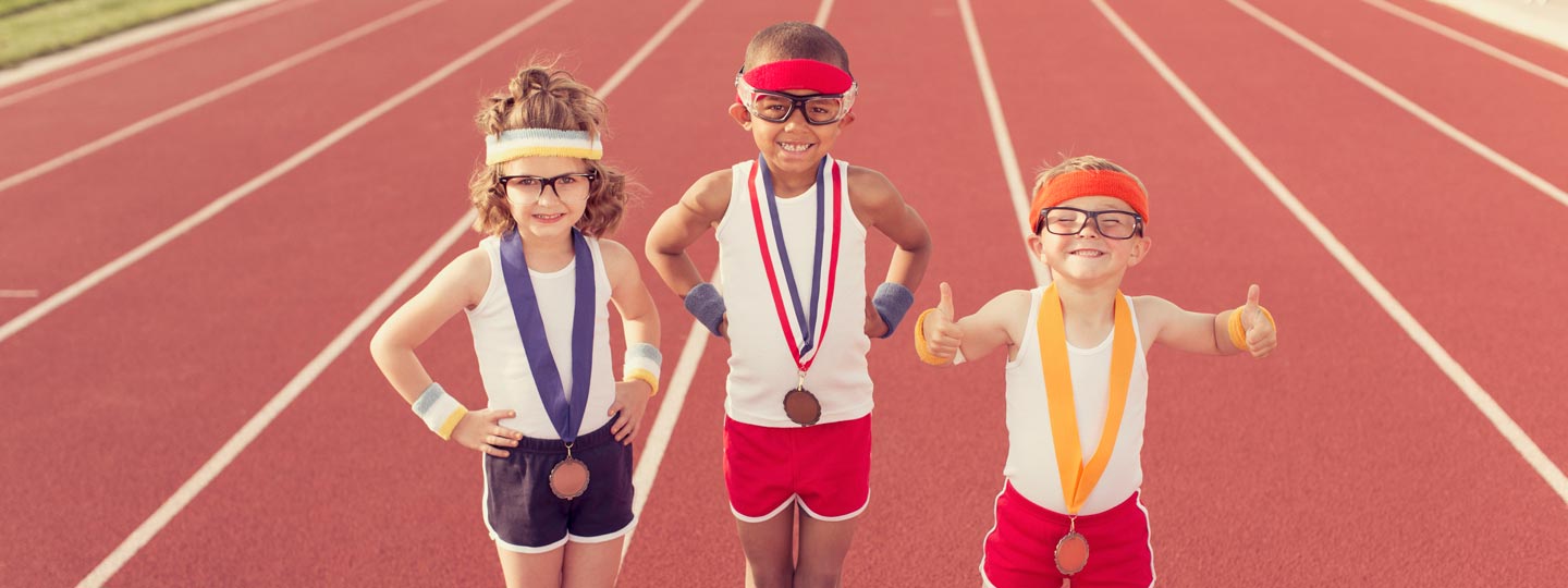 Drei Kinder in Sport-Outfit und mit Medaillen um den Hals stehen auf einer Tartanbahn und lächeln stolz.
