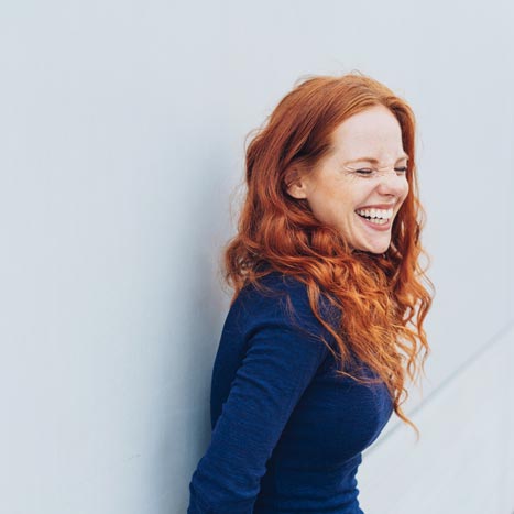 Eine junge, rothaarige Frau steht lachend an einer Wand.